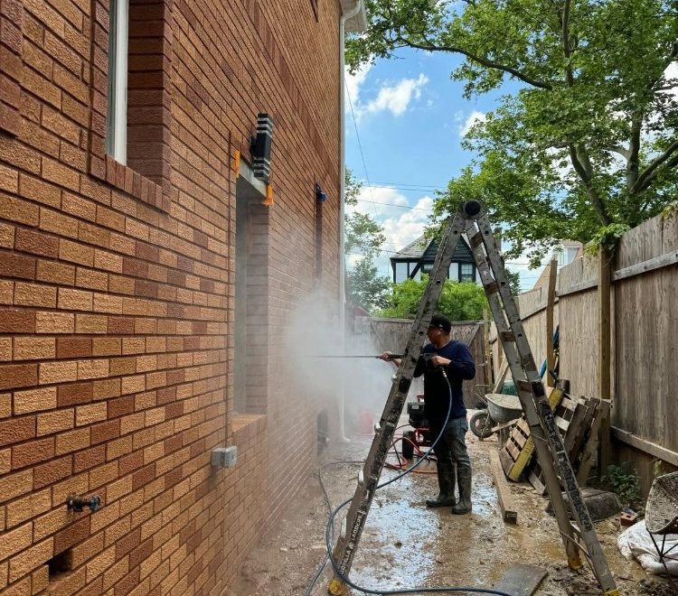 Worker pressure washing a brick wall on a residential property, with a ladder and tools in view, surrounded by greenery.