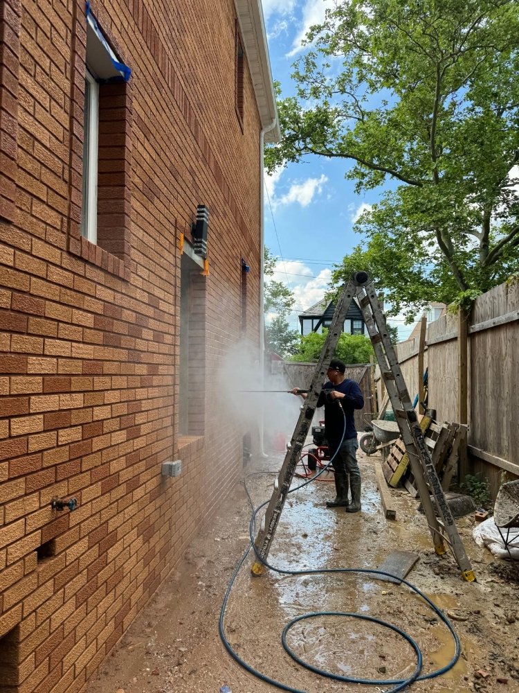 Worker pressure washing a brick wall on a residential property, with a ladder and tools in view, surrounded by greenery.