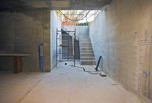 Unfinished basement under construction with concrete walls and stairway, showing early-stage waterproofing preparation.