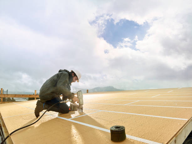 Contractor working on a flat roof, installing BUR layers with a power tool under a cloudy sky.
