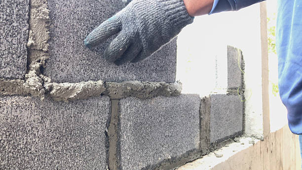 Worker building a concrete block wall using mortar, wearing gloves for safety.